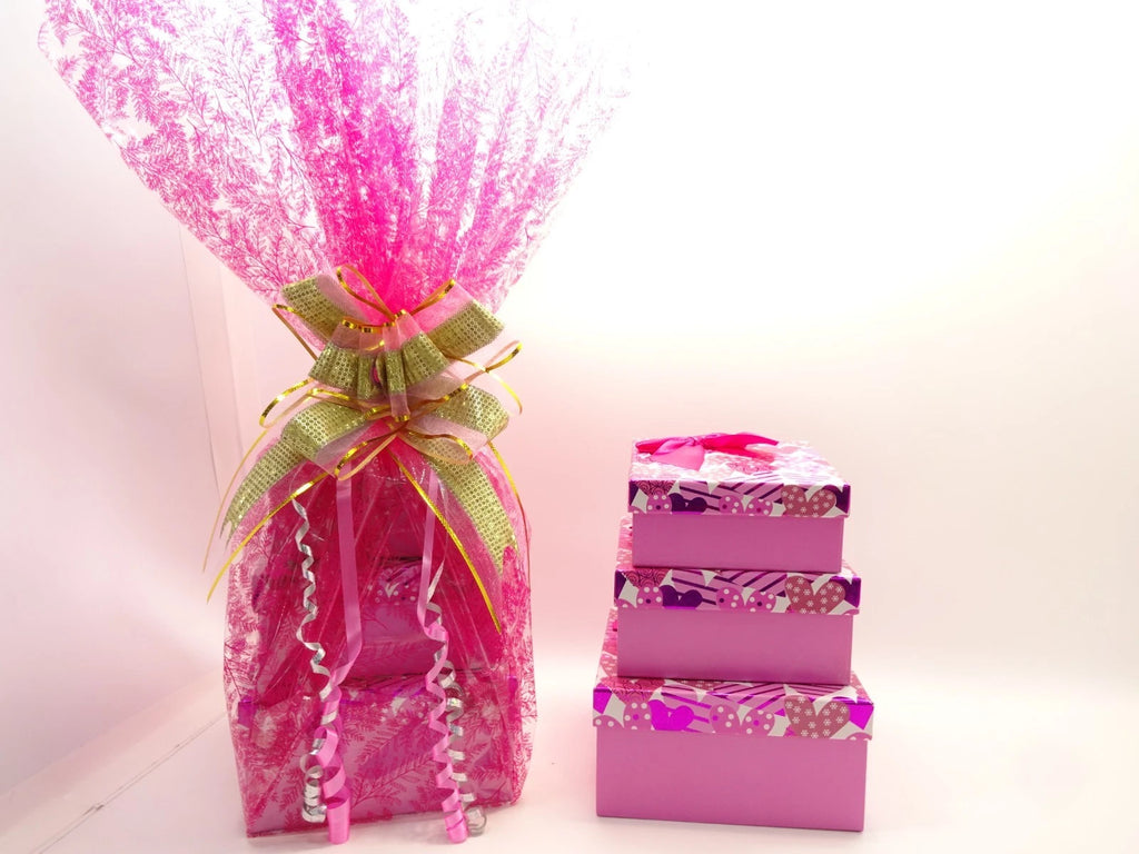 Three pink heart-themed gift boxes stacked next to wrapped hamper with satin bows
